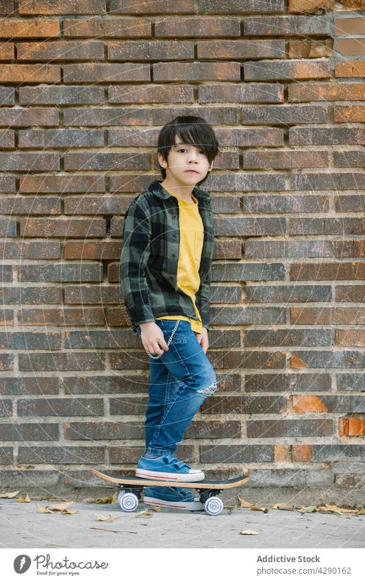 Kleiner Junge steht mit Skateboard auf Backsteinmauer Hintergrund Kind Straße Kindheit Hobby ethnisch sportlich Skateboarderin Streetstyle sorgenfrei