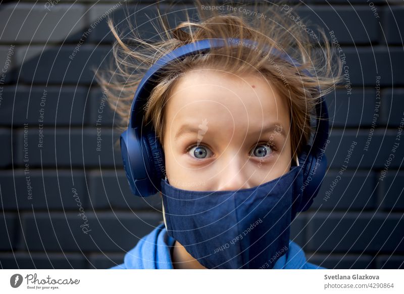 Lustiges Porträt eines blonden, kaukasischen Jungen, dessen Haare aufstehen und im Wind wehen. Schaut in die Kamera.gekleidet in einer schützenden medizinischen Maske und blaue Kopfhörer