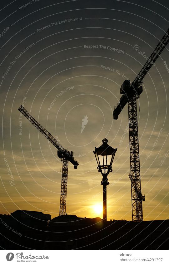 Zwei Baukräne und eine altertümliche Straßenlampe im abendlichen Gegenlicht / Bauplatz / Städteplanung Sonnenuntergang Bauwerk Abendstimmung Architektur golden