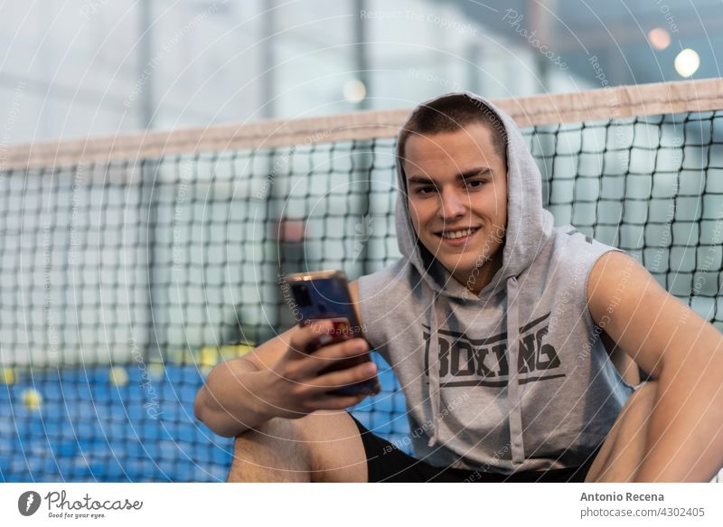 Junger Mann mit urbanen Stil nimmt eine Pause mit dem Telefon Züge nach dem Spielen Paddle-Tennis auf einem Platz im Freien jung Training Paddeltennis Padel