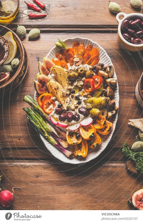 Teller mit frisch zubereitetem Hummus, gegrilltem Gemüse und einer Vielzahl von Nüssen und Samen als Belag. Gesundes veganes Essen. Mittagessen oder Snack auf rustikalem Holzuntergrund. Ansicht von oben