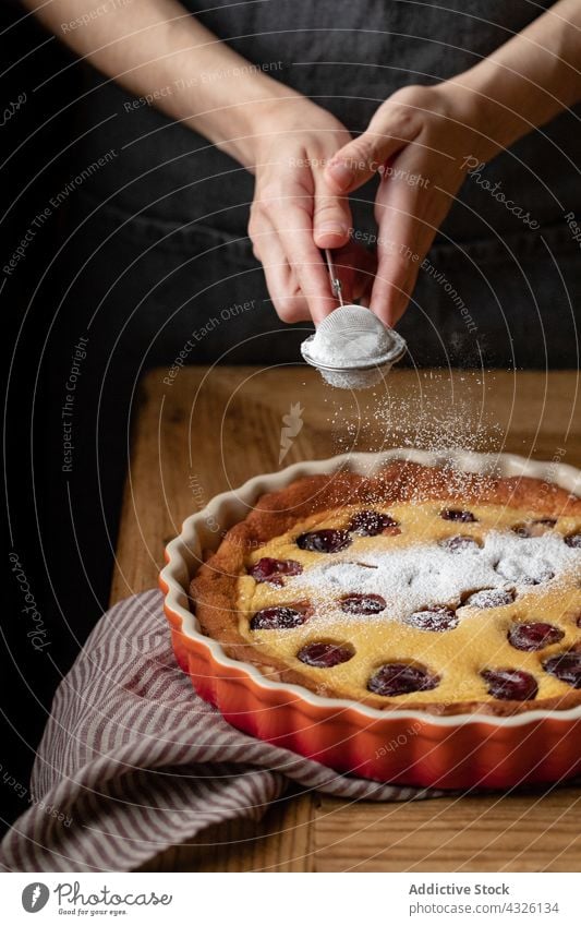 Kropfenkoch gibt Puderzucker auf Kirsch-Clafoutis Pasteten Koch Kirsche hinzufügen Zucker Pulver lecker selbstgemacht Person Schürze gebacken hölzern Tisch