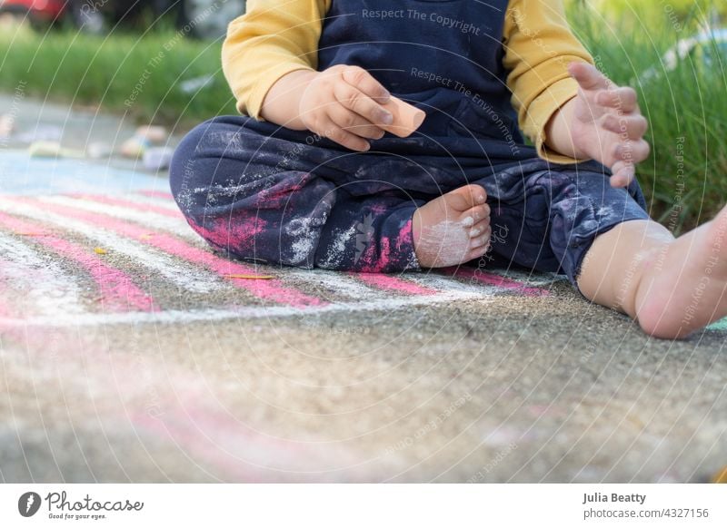 13 Monate altes Baby hält ein Stück Bürgersteig Kreide, Kleinkind sitzt auf einer Zeichnung der amerikanischen Flagge und hat Kreidestaub auf Hose und Körper