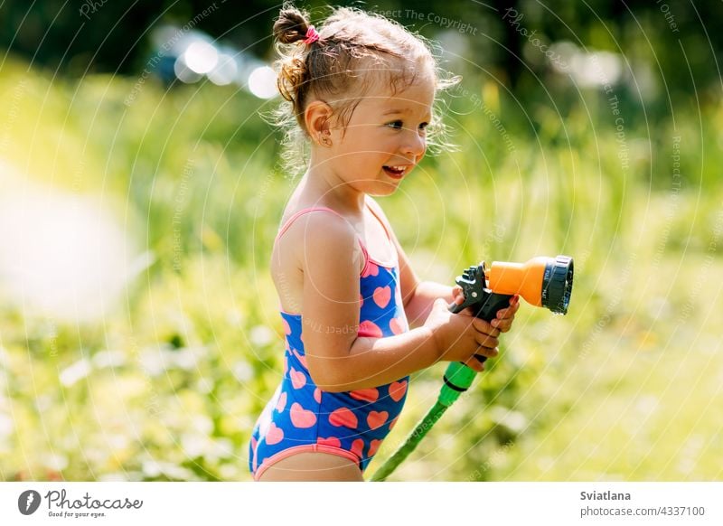 Charmantes Baby Mädchen wässert Pflanzen mit einem Schlauch im Garten im Hinterhof des Hauses an einem sonnigen Sommertag Kind wenig Spray Kindheit Wasser