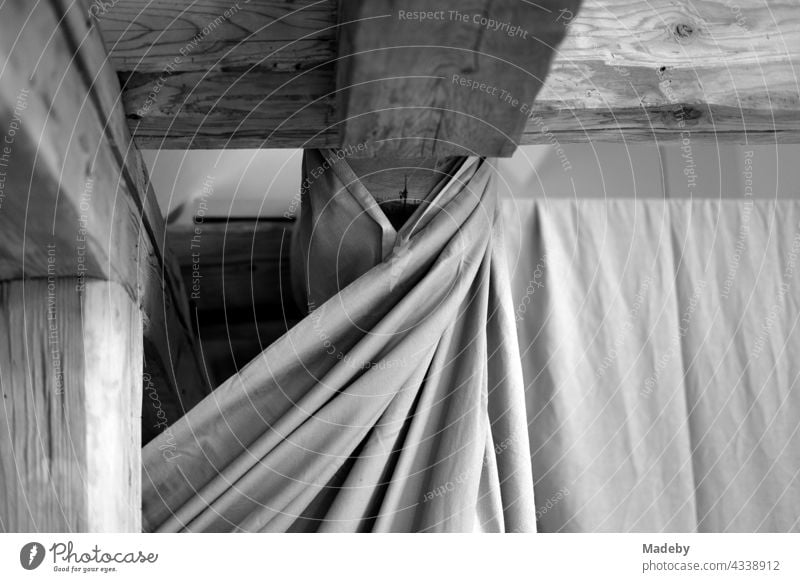 Massive alte Dachbalken aus Holz unter dem Dach und ein Vorhang mit Falten in einem renovierten Bauernhaus auf einem Bauernhof in Rudersau bei Rottenbuch in Oberbayern, fotografiert in traditionellem Schwarzweiß