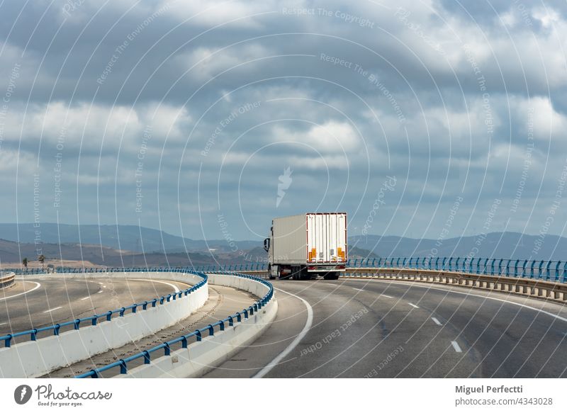 LKW mit Metallsattelauflieger, weit verbreitet in der Textil- und Automobilindustrie, fährt über eine Brücke mit bewölktem Himmel. Lastwagen Anhänger Straße