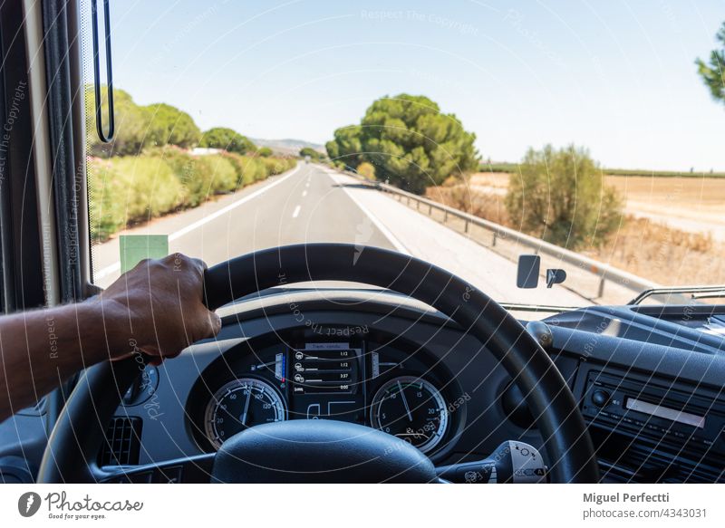 Blick, den ein Lkw-Fahrer von seinem Arbeitsplatz aus hat, mit einer Hand das Lenkrad umgreifend, das Armaturenbrett mit den Auftragsinformationen und die Straße im Hintergrund.