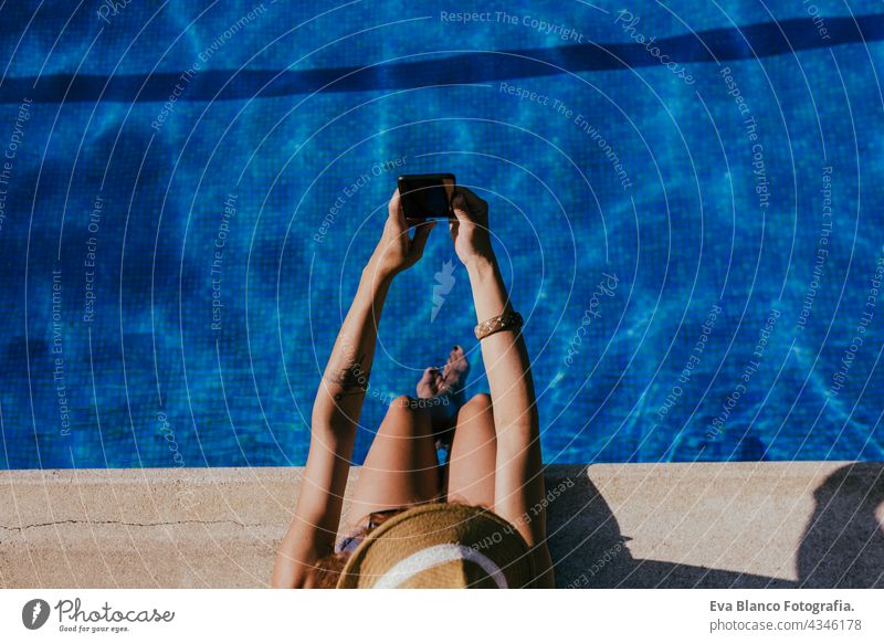 Draufsicht auf junge kaukasische Frau, die am Pool sitzt und ihr Handy benutzt, um ein Selfie zu machen. Sommerzeit, Urlaub und Lebensstil Technik & Technologie