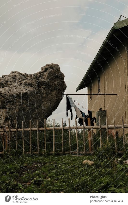 Zwischen Fels und Haus ist eine Wäscheleine mit Wäsche zum Trocknen gespannt. Zaun Garten Kleidung Wiese Rasen düster Fenster Dach grau grün gelb Himmel Wolken