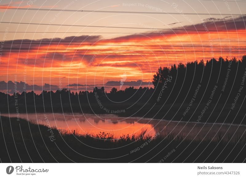 Sonnenuntergang über dem Fluss. Rote orange Wolken. Schwarzer Wald Silhouette über den Fluss. Elektrische Stromleitung Drähte in der Luft. Reflexion von Wolken und Schilf in Fluss macht es orange