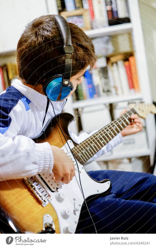 Kind, das zu Hause E-Gitarre spielt, mit einem Kopfhörer und einer unscharfen Bibliothek im Hintergrund, selektiver Fokus elektrisch Stromgitarre Junge Kinder