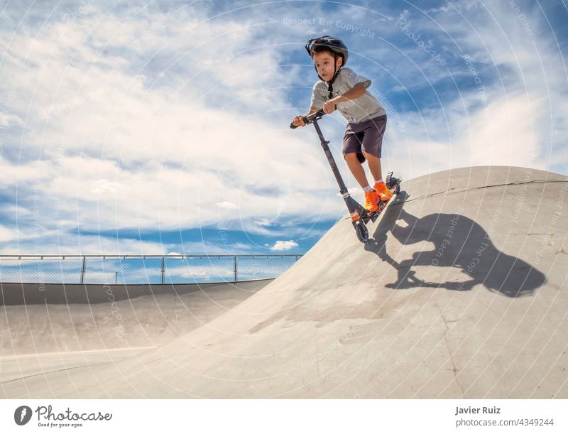 Kind mit Helm fährt an einem sonnigen Sommertag mit einem Roller in einem Skatepark eine Rampe hinunter Tretroller Schlittschuh Skater Junge Skateplatz