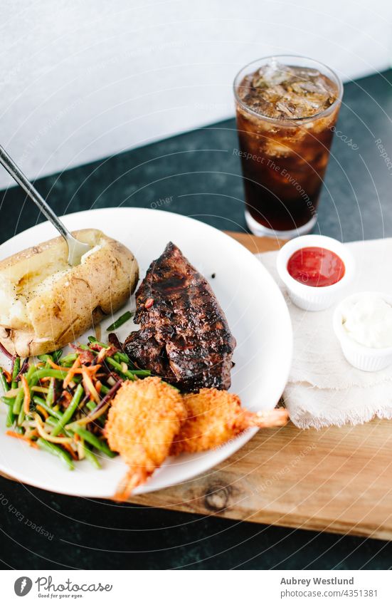 Surf and Turf Steak, Garnelen und Kartoffelessen mit einem Soda vor einem schwarz-weißen Hintergrund angus Ofenkartoffel Barbecue Rindfleisch Holzplatte paniert