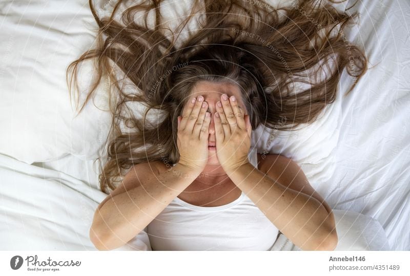 Junge Frau, die im Bett liegt und leidet, müde Frau, die ihr Gesicht mit den Händen bedeckt, kann nicht schlafen und fühlt sich erschöpft auf weißen Laken im Schlafzimmer