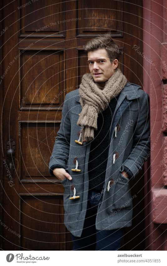 junger Mann mit Dufflecoat wartet im Hauseingang Junger Mann warten Haustür Winter dufflecoat Mantel winterjacke Schal blond stehen anlehnen Blick in die Kamera