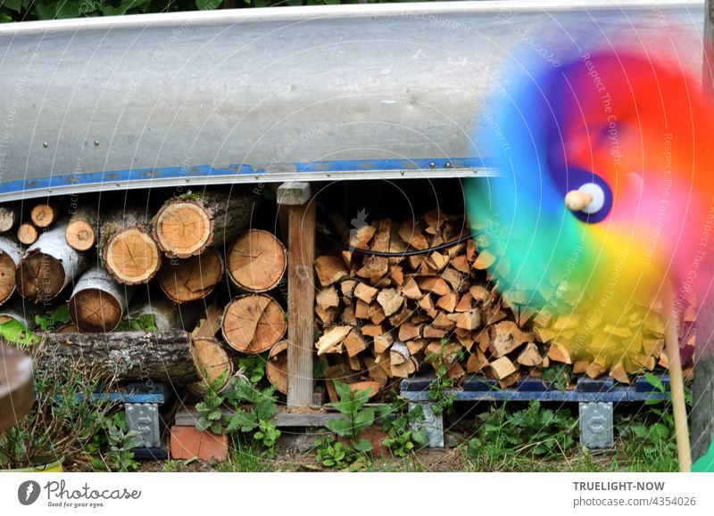 Brennholz für die Feuerschale liegt im Garten auf Paletten gestapelt und geschützt unter einem Kanu und ein buntes Windrad dreht sich schnell im Wind Feuerholz