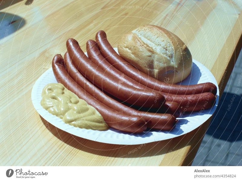 6 Wiener Würstchen aus Wild liegen mit einem Brötchen und Senf auf einem Pappteller, der auf einem Biertisch aus Holz steht. Es ist sonnig. Analoges Bild, 35mm. Körnung.