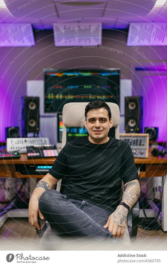 Tonmann mit Tattoos im Musikstudio mit Neonlicht Ingenieur Klang selbstbewusst Beine gekreuzt professionell Atelier Mann Porträt neonfarbig selbstsicher Lächeln