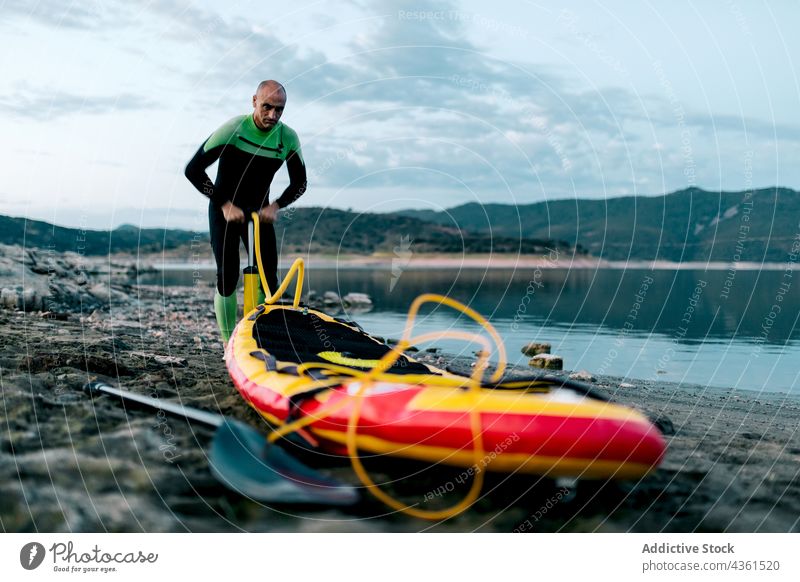 Mann pumpt Paddleboard vor dem Surfen auf Paddelbrett Pumpe SUP Zusatzplatine Surfer vorbereiten Meeresufer Neoprenanzug Strand männlich Surfbrett Sport