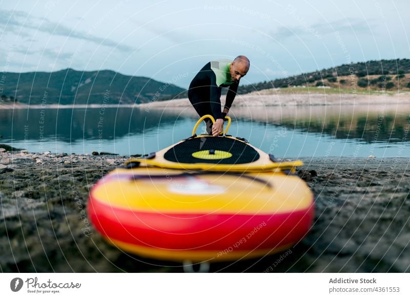 Mann pumpt Paddleboard vor dem Surfen auf Paddelbrett Pumpe SUP Zusatzplatine Surfer vorbereiten Meeresufer Neoprenanzug Strand männlich Surfbrett Sport