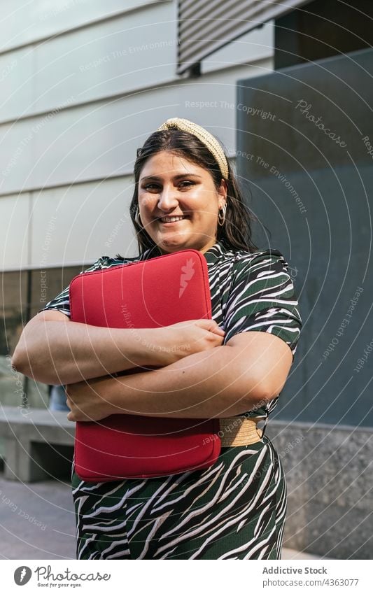 Glückliche Frau mit Laptop-Tasche in der Nähe eines städtischen Gebäudes Lächeln Fall urban modern kurvenreich Stil Farbe Übergröße Streifen Geometrie jung