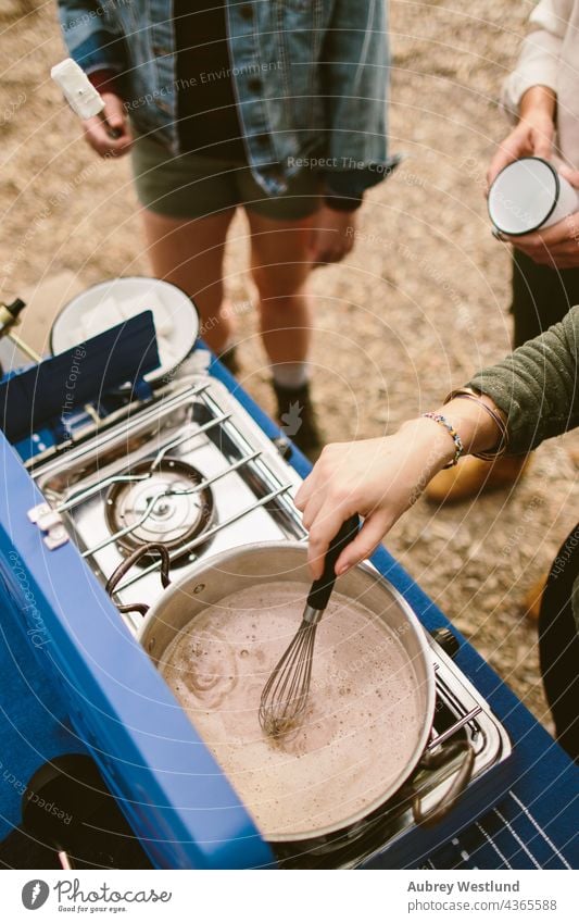 Frau rührt heiße Schokolade auf einem Campingkocher im Freien blau Küchenchef Gemeinschaft Koch Essen zubereiten Kochgeschirr kulinarisch flache Verlegung