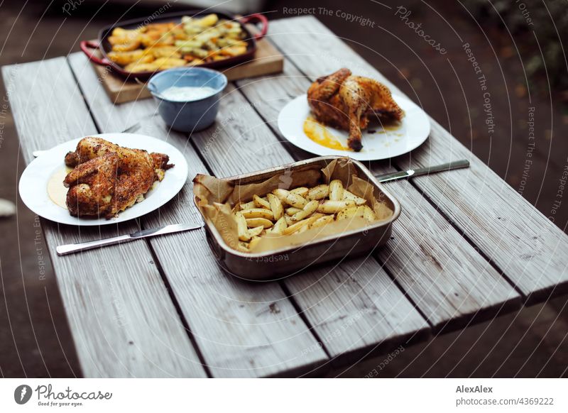 Abendbrottisch aus Holz mit Grillhähnchen, Grillkartoffel und Grillgemüse, draussen. Abendessen Tisch Kartoffeln Hähnchen Broiler BBQ Kartoffelspalten Teller