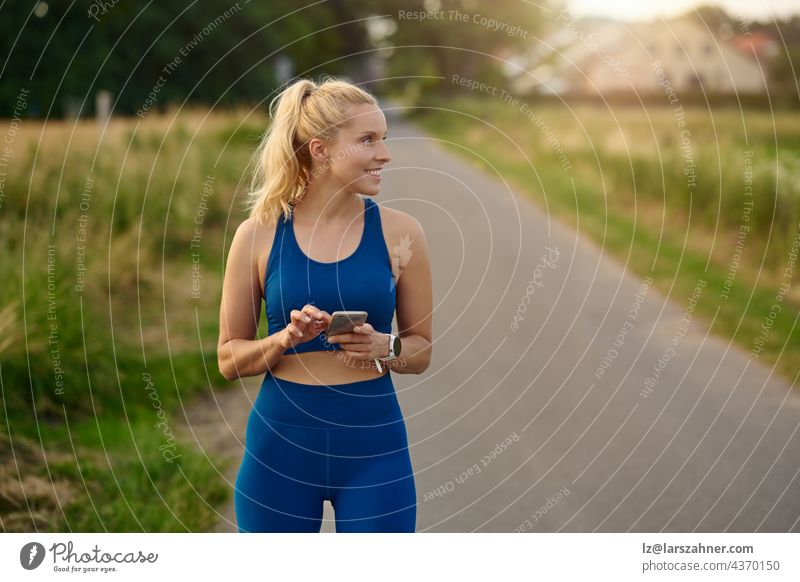 Fitte, sportliche Frau, die beim Joggen auf einem ländlichen Wanderweg ihr Handy konsultiert und dabei mit einem zufriedenen Lächeln zur Seite schaut passen