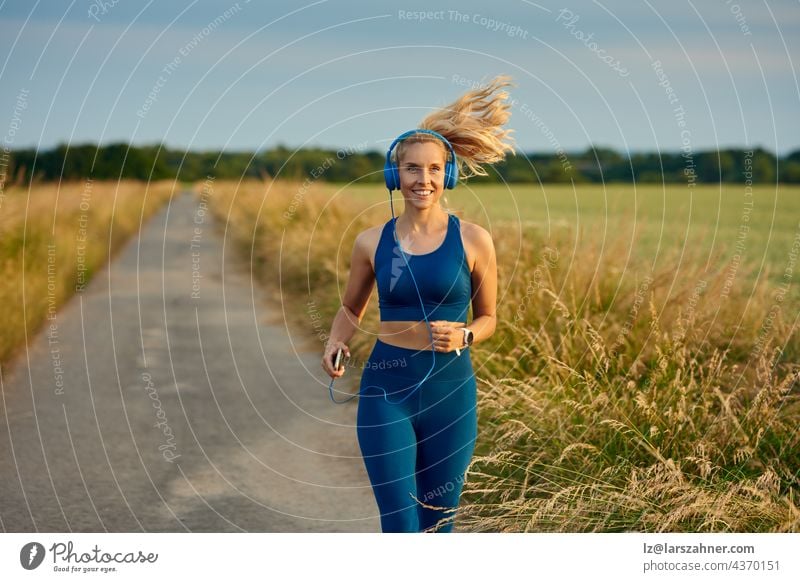 Eine fitte, lebhafte junge Frau, die auf einem Fußweg in einem offenen Feld joggt, nähert sich der Kamera mit einem glücklichen Lächeln und einem nach hinten fliegenden Pferdeschwanz in einem gesunden, aktiven Lebensstilkonzept.