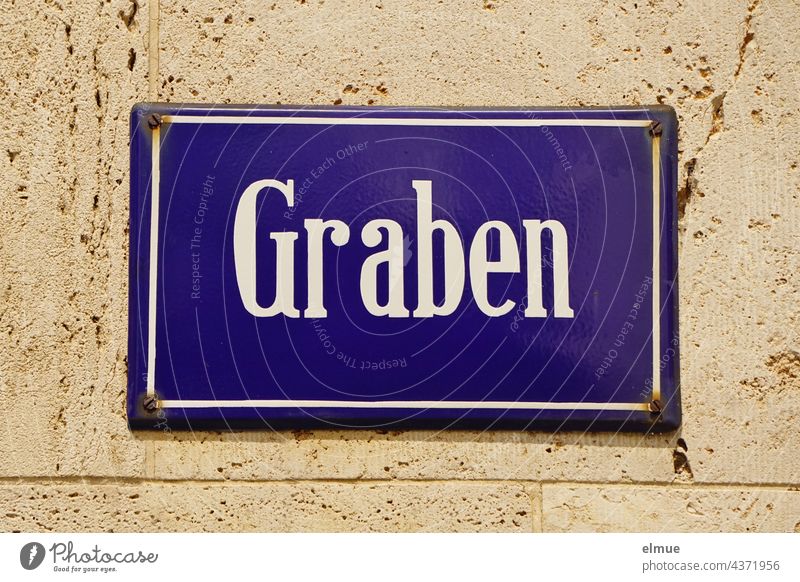 blaues Straßennamenschild  Graben an einer hellen Wand / Adresse / Straßenverzeichnis wohnen Hausmauer Orientierung Schild Straßenbezeichnung Symbol