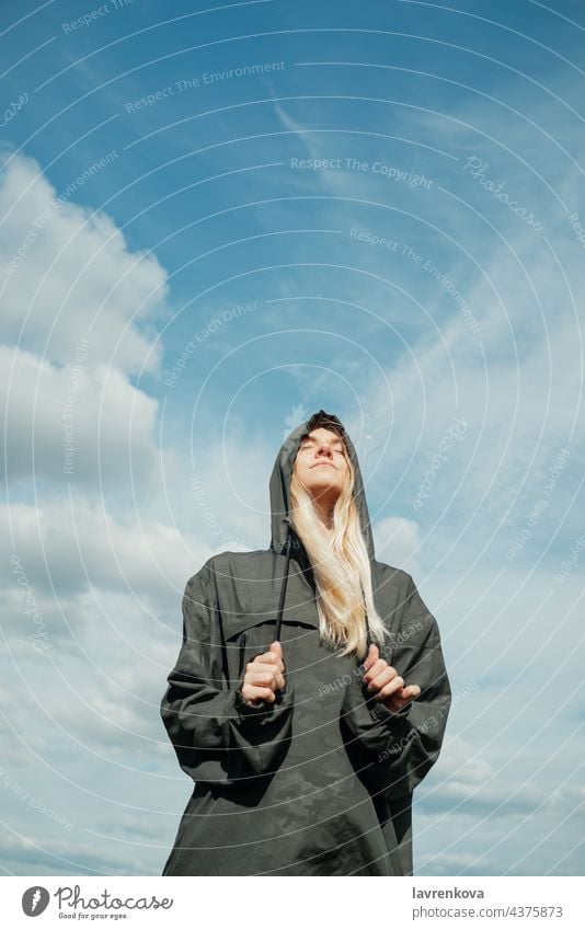 Junge blonde Frau in khakifarbenem Hemd mit Kapuze steht mit geschlossenen Augen vor dem blauen Himmel Augen geschlossen khakigrün hübsch vertikal Landschaft