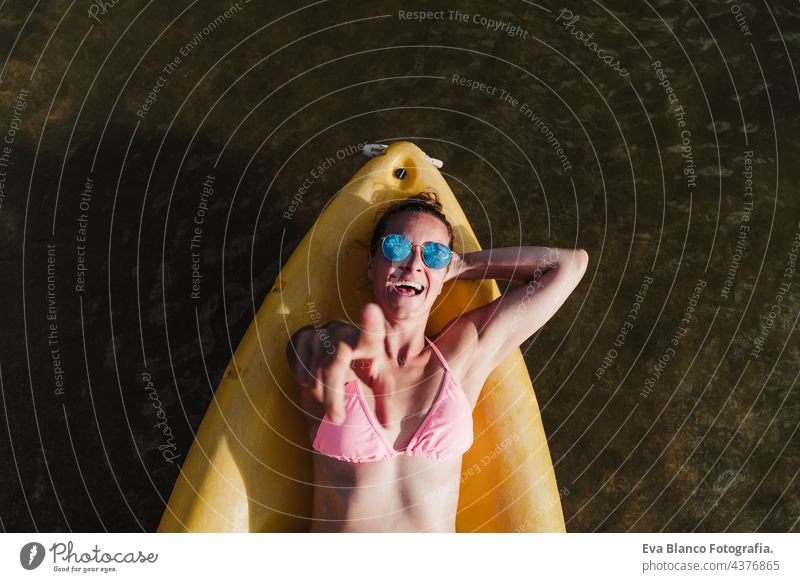 Draufsicht auf glückliche kaukasische Frau, die auf einem gelben Kanu im See liegt, während eines sonnigen Tages, der mit dem Finger auf die Kamera zeigt. Sport, Abenteuer und Natur. Frau in Badehose und Sonnenbrille