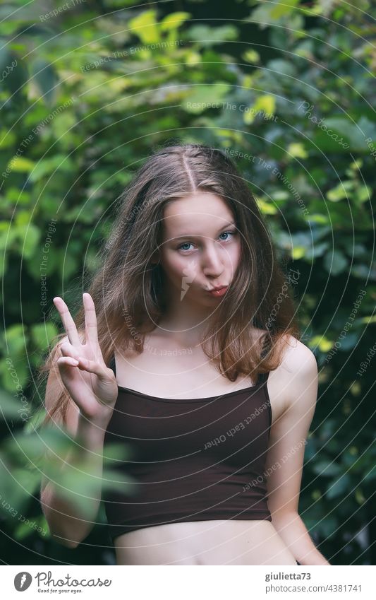 Sommerliches Porträt eines langhaarigen Teenager Mädchens, dass einen Kussmund macht und das Victory-Zeichen zeigt Vorderansicht jung blond verschmitzter Blick