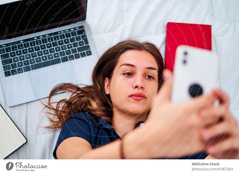 Studentin macht Selfie mit Smartphone Frau Schüler zu Hause online Apparatur Bett Pause abgelegen jung fotografieren Gerät Laptop Mobile benutzend Internet