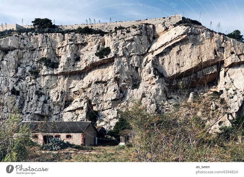 Felswand mit Haus Gestein klein Himmel Licht trocken Gestrüpp massiv feindlich grau Gesteinsschichten Außenaufnahme Farbfoto Natur Felsen Tag Berge u. Gebirge