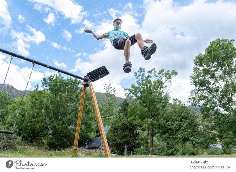 Ein junger Mann, der eine Hygienemaske trägt, springt von einer Schaukel auf einem Spielplatz in das leere Wasser. springend pendeln fliegen Gesichtsmaske