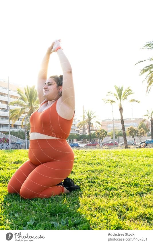 Plus Größe ethnischen Sportlerin Stretching Arme auf Rasen im Sonnenlicht Athlet Arme hochgezogen Dehnung Training Übung Übergröße Frau Aufwärmen mollig Wiese