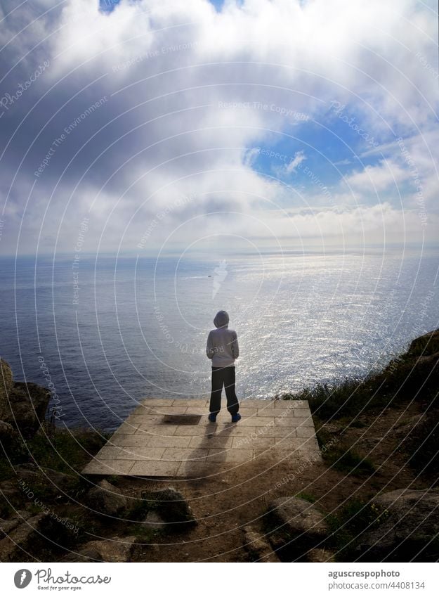 Unbekannter Junge mit Kapuzenpulli auf dem Rücken, der vom Rand der Küstenlinie auf eine Klippe in Finisterre, La Coruña, Spanien, blickt. Im Hintergrund ist der Horizont des Meeres und ein Himmel mit Wolken.