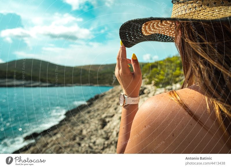 Seitenansicht eines nicht wiederzuerkennenden brünetten Mädchens, das sich im Sommer an ihrem Heuhut festhält. Stehend in der Nähe der Küste des adriatischen Meeres, Konzepte von Reisen, Sommer und Urlaub