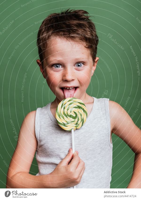 Lustiger Junge leckt süßen Lutscher am Stiel im Studio Kind Lollipop Zunge Bonbon lutschen Gesicht machen lecker Spaß haben lustig geschmackvoll Leckerbissen