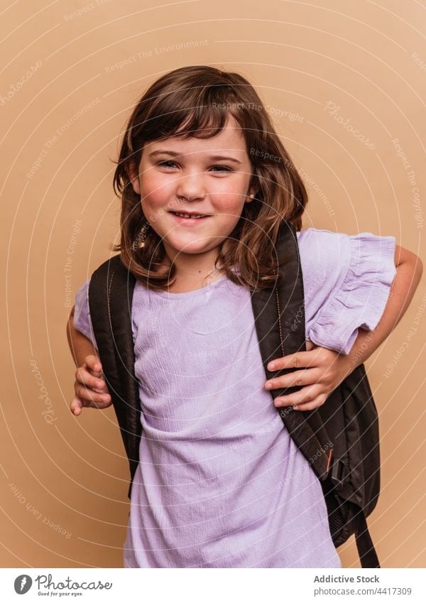 Charmantes Mädchen mit Rucksack auf braunem Hintergrund Schulmädchen Entengesicht Kind Atelier Gesicht machen Schulkind Pupille Vorschein niedlich bezaubernd