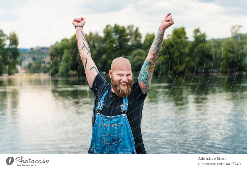 Fröhlicher Mann mit erhobenen Händen am See Lächeln Natur Sommer Hipster Tattoo Baum Vollbart Wasser Spaß heiter jung spielerisch brutal Glück lässig männlich