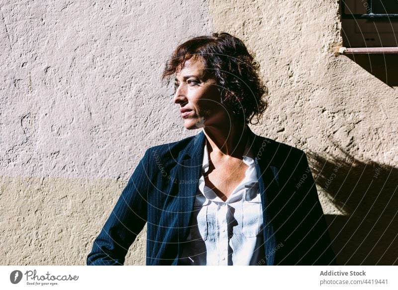 Verträumte Frau in formeller Kleidung steht in der Nähe einer Steinmauer verträumt Sonnenlicht Starrer Blick nachdenklich Schatten Persönlichkeit elegant