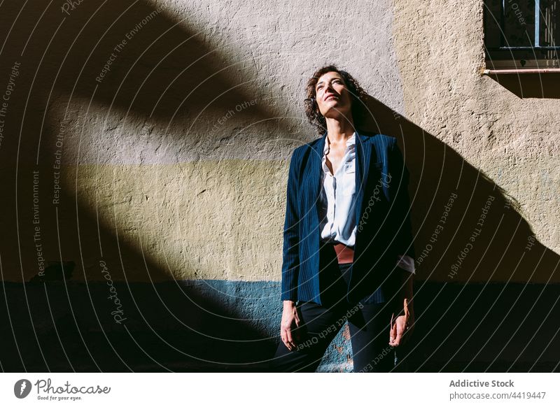 Verträumte Frau in formeller Kleidung steht in der Nähe einer Steinmauer verträumt Sonnenlicht Starrer Blick nachdenklich Schatten Persönlichkeit elegant