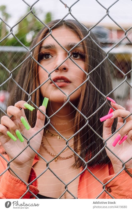 Junge Frau hinter Metallgitter stehend tausendjährig Porträt trendy cool Stil Maniküre lang Raster selbstbewusst charmant feminin Interesse Starrer Blick ernst