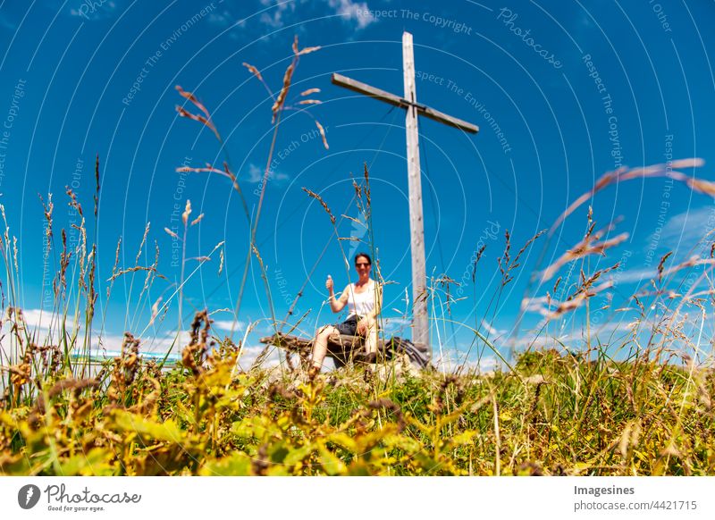 Verschwommen. Gipfelkreuz auf dem Gipfel des Berges in den bayerischen Alpen mit einem Bergsteiger, der die Daumen hochhebt. Angekommen, Siegerpose oben an einem Bergkreuz. Fokus auf Gras.