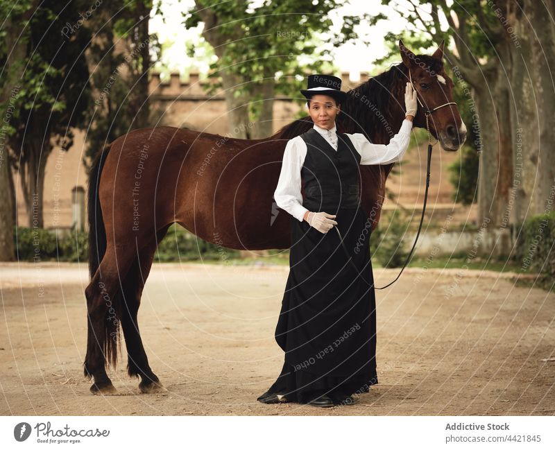 Elegante schwarze Frau mit Pferd im Garten selbstbewusst elegant Jockey Baum Hof Gebäude historisch Landschaft Starrer Blick nachdenken Tier Afroamerikaner