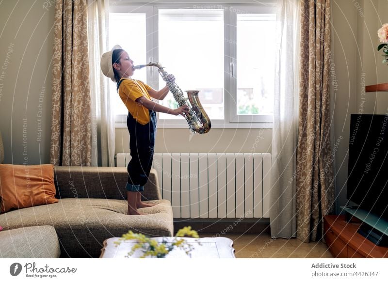 Mädchen spielt Saxophon auf Sofa in Haus Zimmer spielen Musik Kunst klassisch Klang Melodie Raum Kindheit Fenster Liege heimwärts Barfuß ausführen Musiker Hut