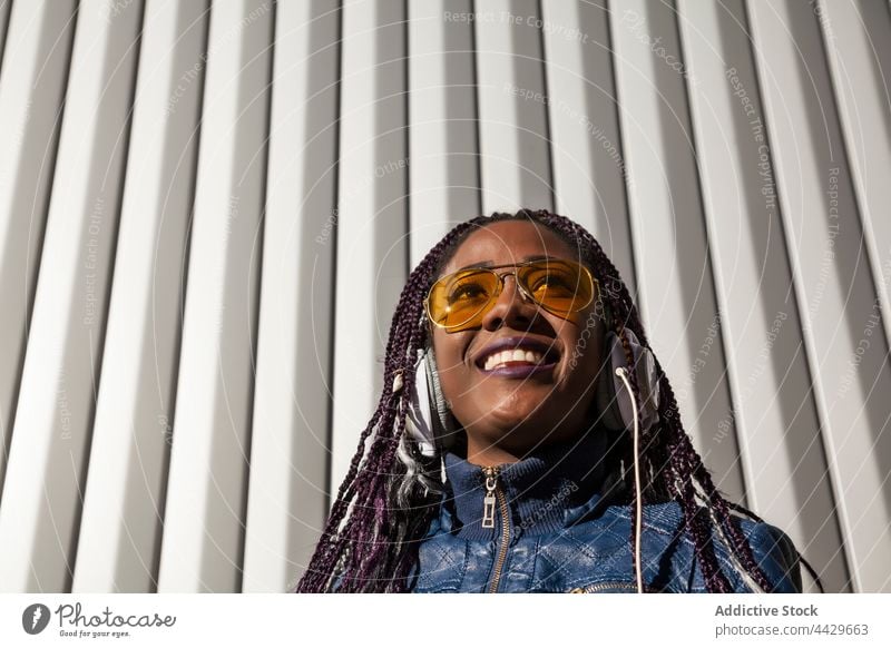 Fröhliche schwarze Frau beim Musikhören Kopfhörer zuhören Stil trendy heiter Kälte Glück genießen Afroamerikaner ethnisch Afro-Look Geflecht Sonnenbrille jung