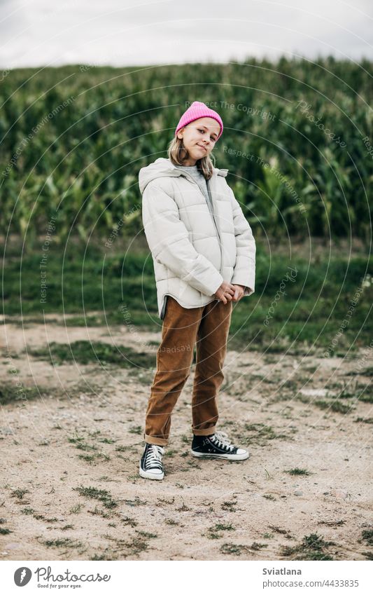 Ein Teenager-Mädchen in einem stilvollen Bild und einer rosa Mütze steht vor dem Hintergrund eines Maisfeldes Herbst gelb Kornfeld Umhang Lachen Lifestyle